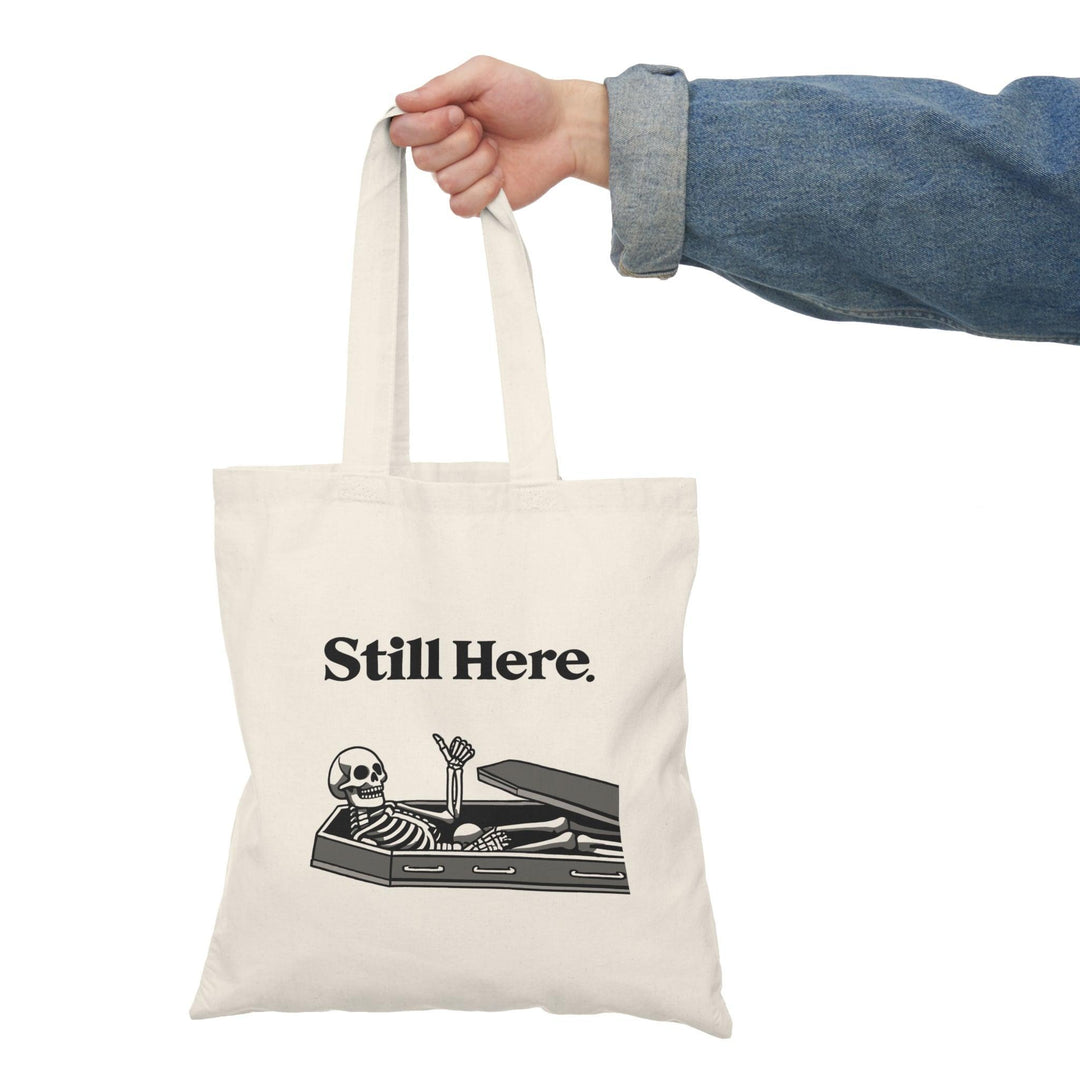 A natural canvas tote bag featuring an illustration of a skeleton lying in a coffin with the text 'Still Here.' The humorous design adds a touch of gothic charm, making it a fun accessory for those who enjoy Halloween-themed or quirky designs