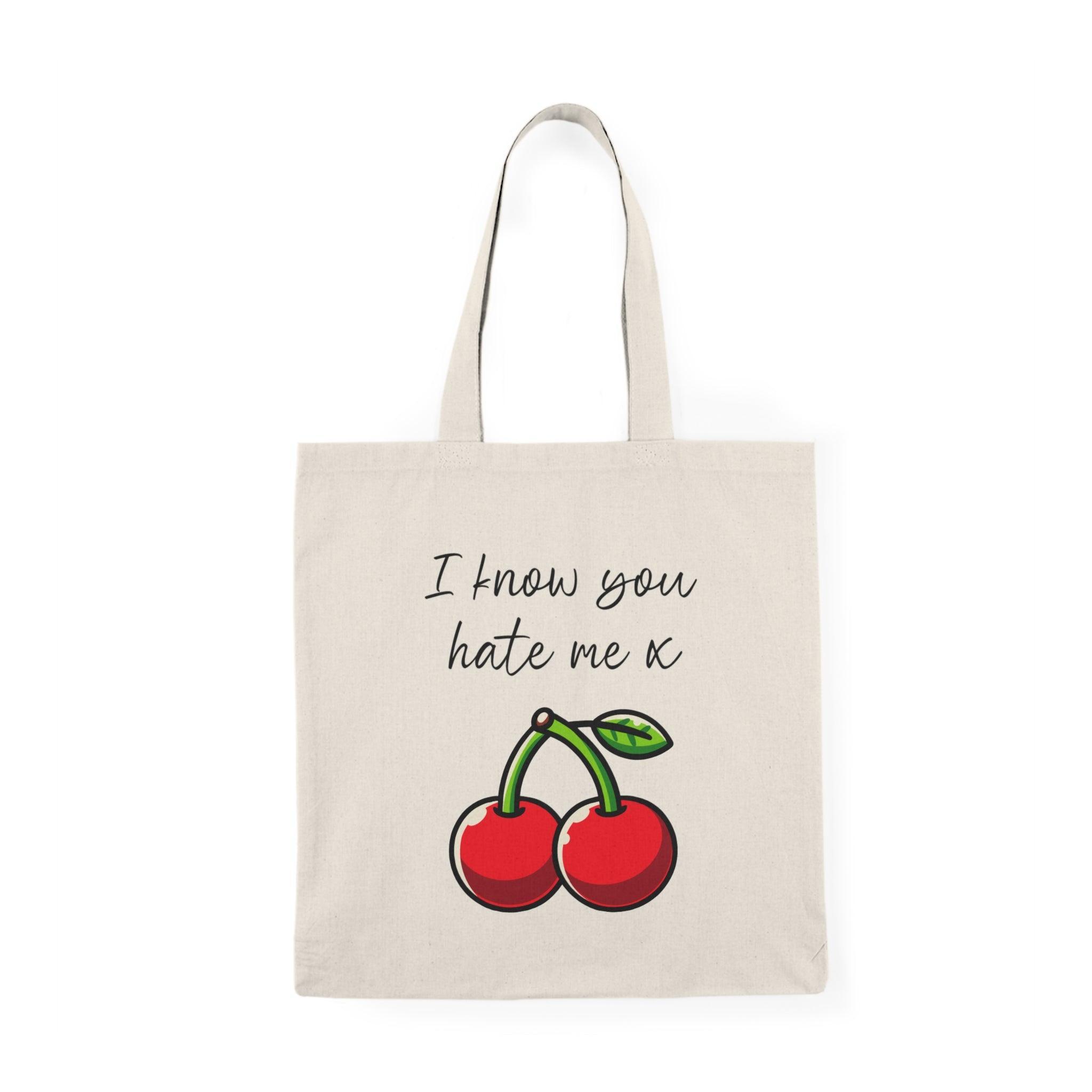 A natural tote bag showcasing a bold design of two cherries, with the text 'I Know You Hate Me' written in a fun, casual font. The combination of the colorful illustration and cheeky message makes this tote a fun accessory for any casual outing