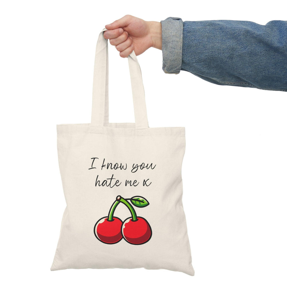 A natural tote bag showcasing a bold design of two cherries, with the text 'I Know You Hate Me' written in a fun, casual font. The combination of the colorful illustration and cheeky message makes this tote a fun accessory for any casual outing