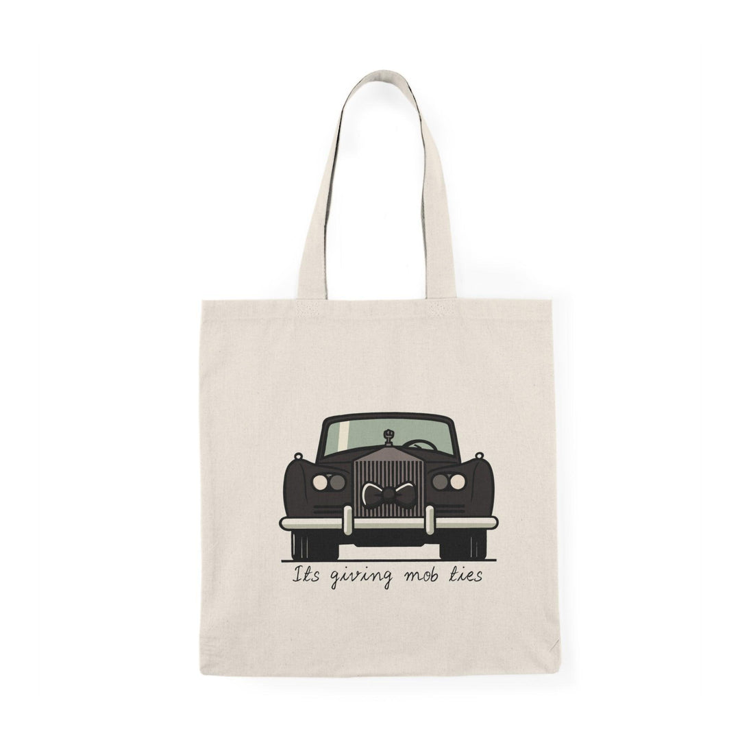 A cream tote bag featuring a vintage car illustration with the phrase 'It's giving mob ties' written underneath. The design captures a retro aesthetic with a touch of humor, making it a unique accessory for car enthusiasts and those with a flair for the dramatic.