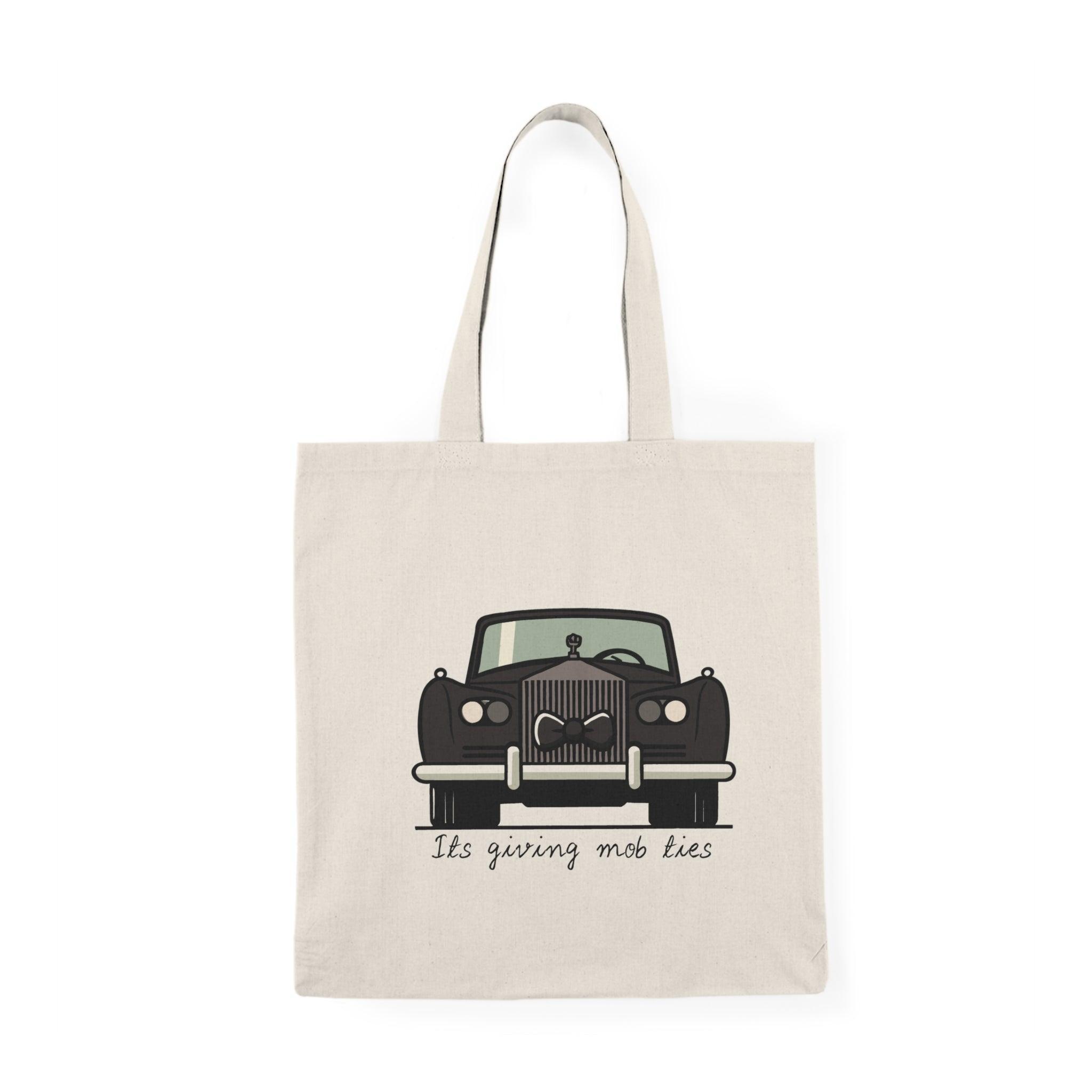 A cream tote bag featuring a vintage car illustration with the phrase 'It's giving mob ties' written underneath. The design captures a retro aesthetic with a touch of humor, making it a unique accessory for car enthusiasts and those with a flair for the dramatic.