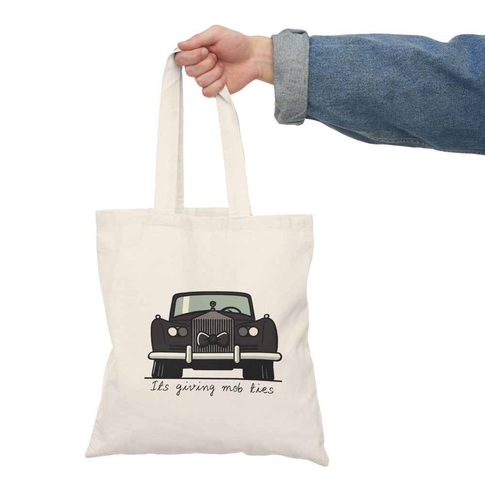 A cream tote bag featuring a vintage car illustration with the phrase 'It's giving mob ties' written underneath. The design captures a retro aesthetic with a touch of humor, making it a unique accessory for car enthusiasts and those with a flair for the dramatic.