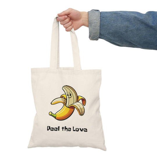 A natural canvas tote bag adorned with a whimsical graphic of a smiling banana character with the phrase 'Peel the Love.' The fun and vibrant design makes this tote a charming choice for fruit lovers and adds a playful element to everyday errands.