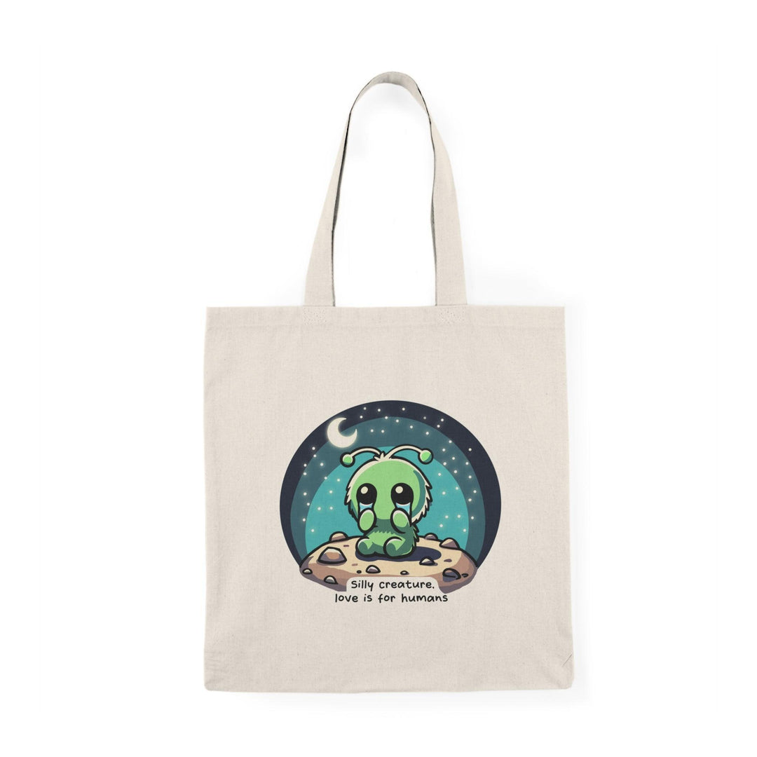 A natural canvas tote bag showcasing a whimsical design of a cute alien character surrounded by stars and the text 'Silly Creature, Love is for Humans.' This fun and colorful graphic adds a touch of humor and charm, making it a delightful accessory for those who enjoy quirky designs