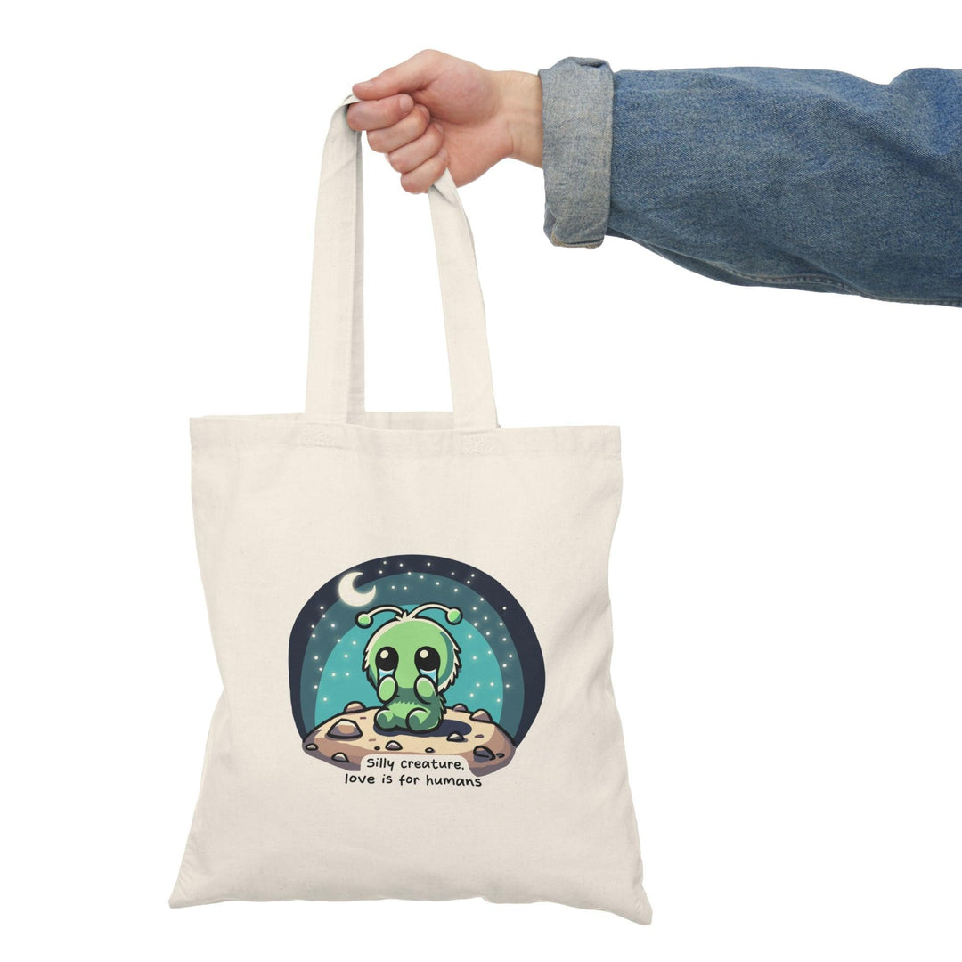 A natural canvas tote bag showcasing a whimsical design of a cute alien character surrounded by stars and the text 'Silly Creature, Love is for Humans.' This fun and colorful graphic adds a touch of humor and charm, making it a delightful accessory for those who enjoy quirky designs