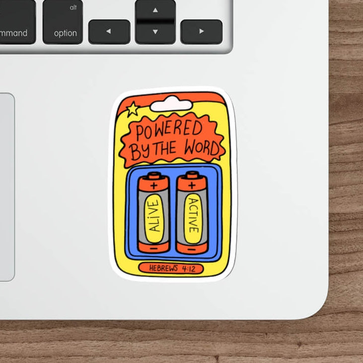 A die-cut sticker featuring two cartoon batteries with the text 'POWERED BY THE WORD' and 'BATTERY LIFE' is low, placed on the corner of a silver laptop on a wooden desk next to a mouse and stylus.