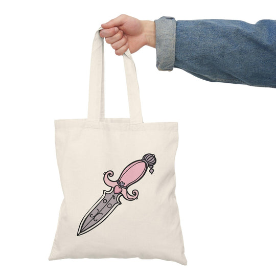 A cream-colored tote bag featuring a bold illustration of a pink dagger with intricate designs on the blade. The eye-catching graphic adds a touch of edgy style, making it a great accessory for those who appreciate unique and artistic designs.