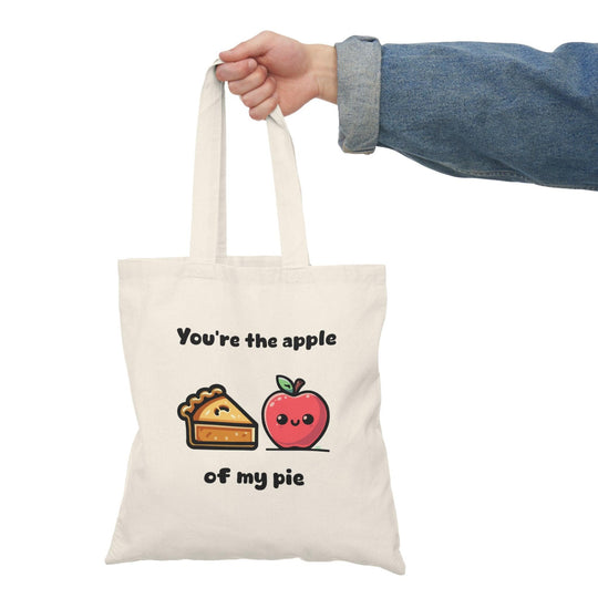A cream tote bag featuring a delightful graphic of an apple pie with apples on either side and the text 'You're the apple of my pie.' This punny design adds a touch of sweetness and charm, making it an ideal gift for food lovers and those with a sweet tooth.