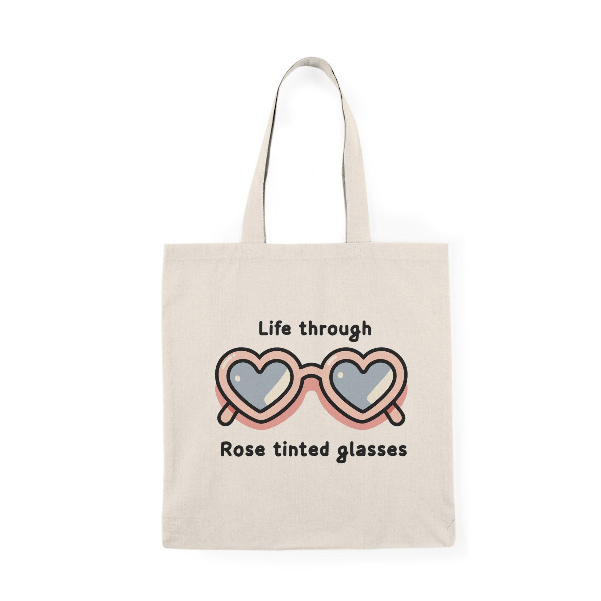 A natural canvas tote bag adorned with an artistic graphic of two overlapping heart shapes, symbolizing rose-tinted glasses. The text 'Life Through Rose Tinted Glasses' is playfully integrated into the design, reflecting a positive outlook and love for life.