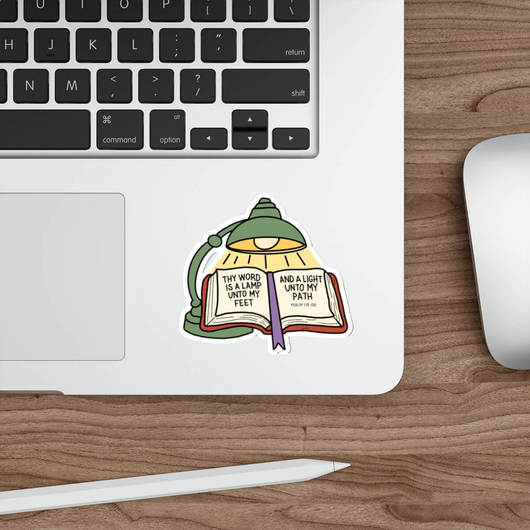 A die-cut sticker featuring a green desk lamp illuminating an open book with a Bible verse printed on the pages, shown applied to the palm rest of a silver laptop on a wooden desk next to a mouse and stylus.