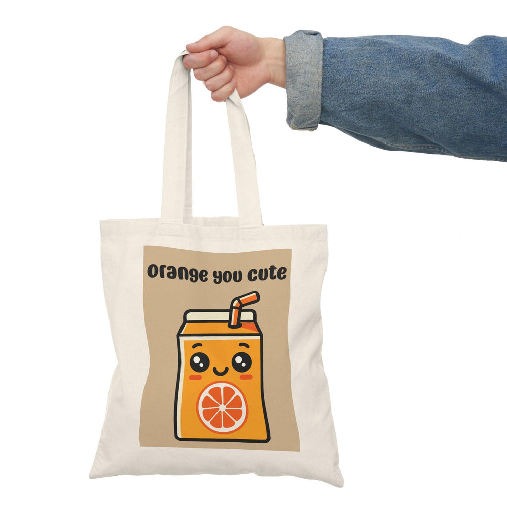 A cream-colored tote bag featuring an adorable graphic of a cartoon-style orange juice carton with a smiling face and the text 'Orange You Cute' in a playful font. This cheerful design adds a fruity flair, perfect for brightening up any casual outfit
