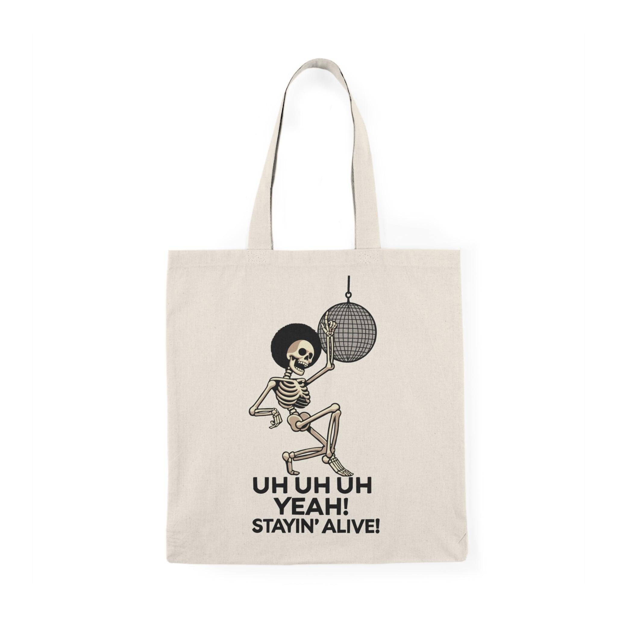 A cream-colored tote bag featuring a fun graphic of a dancing skeleton with the text 'Uh Uh Oh Yeah! Stayin' Alive.' The playful design captures a lighthearted spirit and a classic disco vibe, perfect for music lovers and those who appreciate quirky accessories.