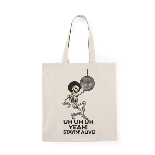 A cream-colored tote bag featuring a fun graphic of a dancing skeleton with the text 'Uh Uh Oh Yeah! Stayin' Alive.' The playful design captures a lighthearted spirit and a classic disco vibe, perfect for music lovers and those who appreciate quirky accessories.