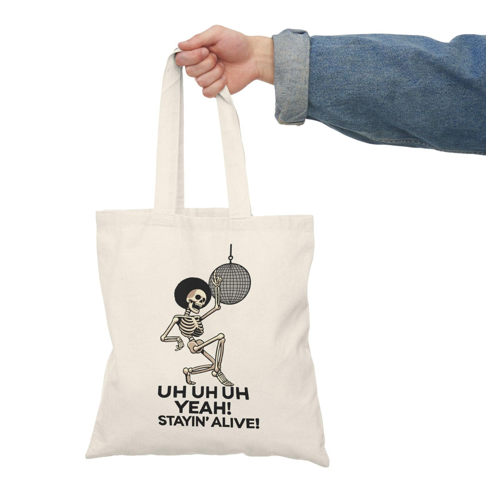 A cream-colored tote bag featuring a fun graphic of a dancing skeleton with the text 'Uh Uh Oh Yeah! Stayin' Alive.' The playful design captures a lighthearted spirit and a classic disco vibe, perfect for music lovers and those who appreciate quirky accessories.
