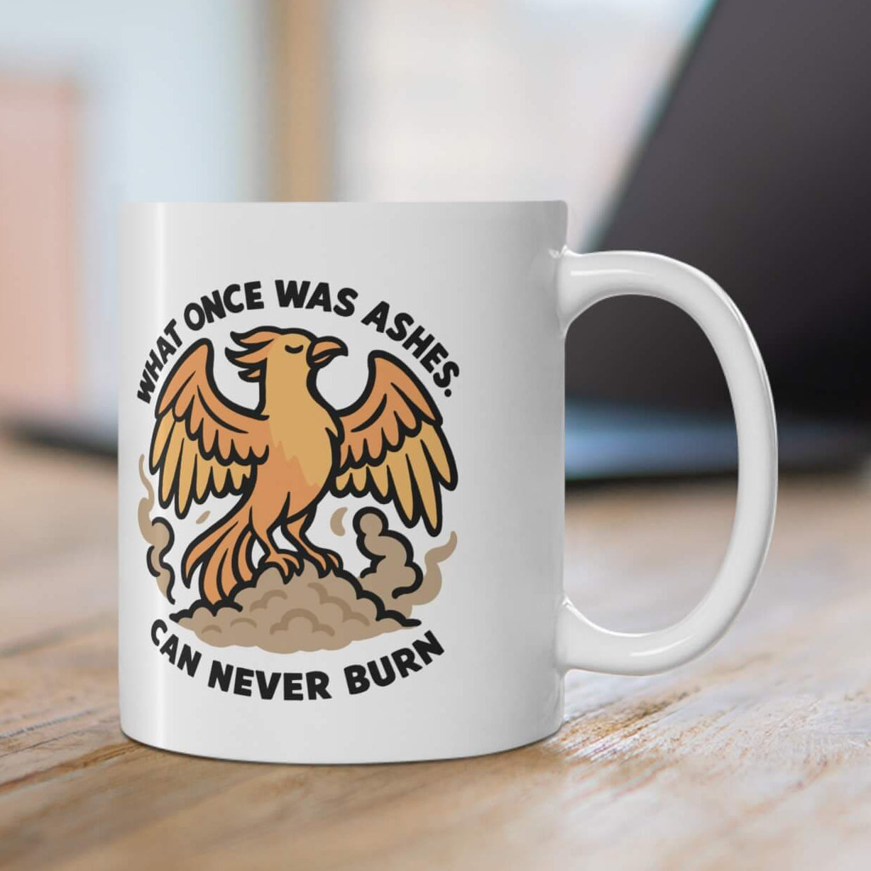 White ceramic mug on wooden table with phoenix rising from smoke and the bold quote “What once was ashes can never burn” in black letters, laptop in background.