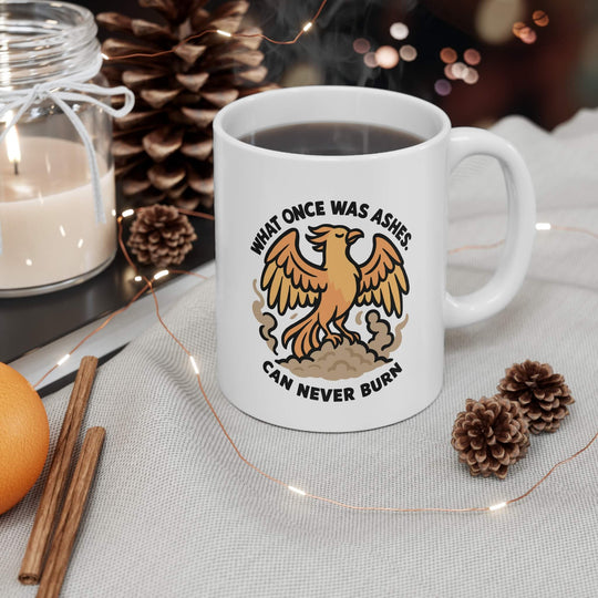 White ceramic mug on wooden table with phoenix rising from smoke and the bold quote “What once was ashes can never burn” in black letters, laptop in background.