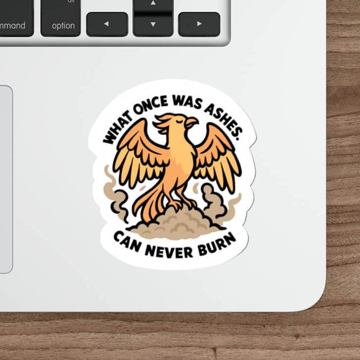 Illustrated phoenix sticker rising from ashes with the quote "What once was ashes can never burn" — shown on a laptop near a keyboard and mouse.

