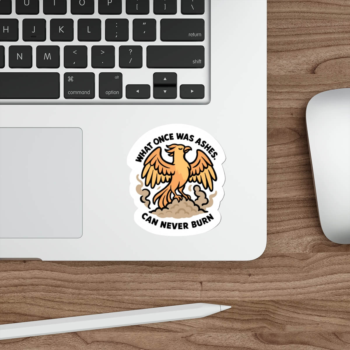 Illustrated phoenix sticker rising from ashes with the quote "What once was ashes can never burn" — shown on a laptop near a keyboard and mouse.

