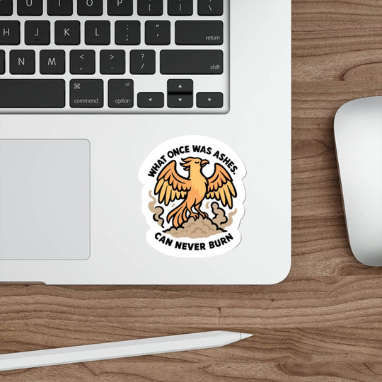 Illustrated phoenix sticker rising from ashes with the quote "What once was ashes can never burn" — shown on a laptop near a keyboard and mouse.

