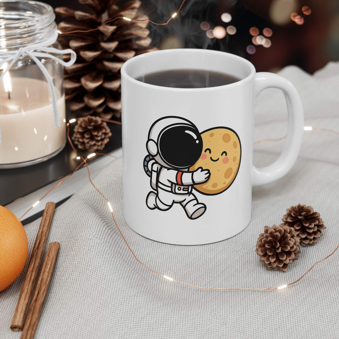 White ceramic mug with a cute cartoon astronaut hugging a smiling moon. Styled with pinecones and cozy winter decor.
