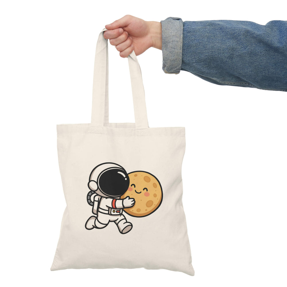 Natural cotton tote bag with a cute cartoon astronaut hugging a smiling moon, held by a hand in a denim jacket.