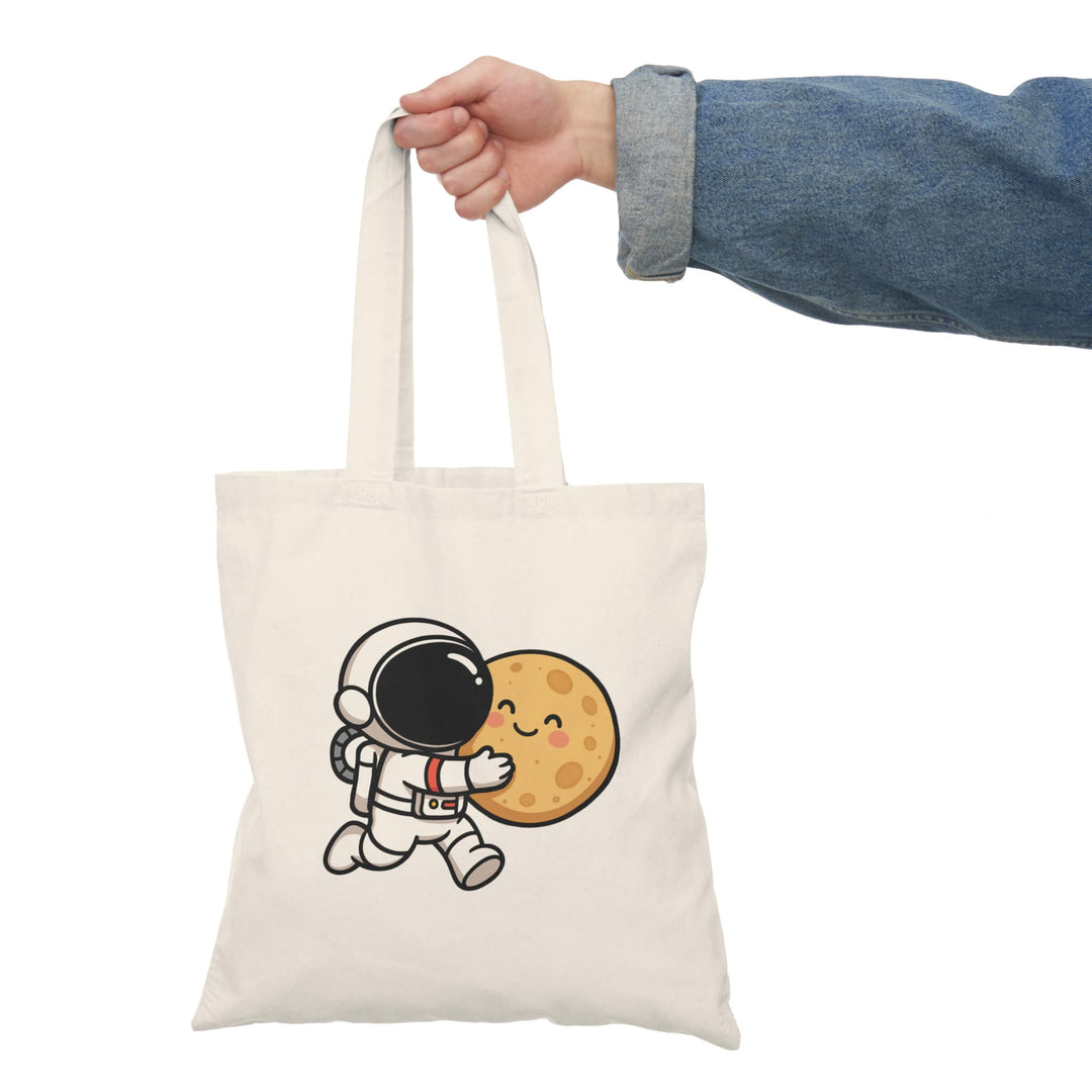 Natural cotton tote bag with a cute cartoon astronaut hugging a smiling moon, held by a hand in a denim jacket.
