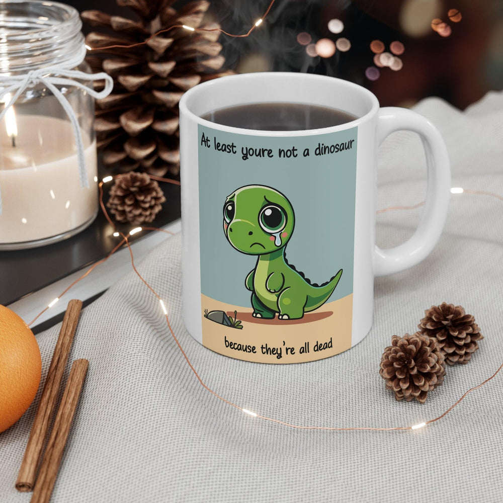 White ceramic mug with a cute dinosaur illustration and the humorous text 'At least you’re not a dinosaur,' adding fun to any morning routine.