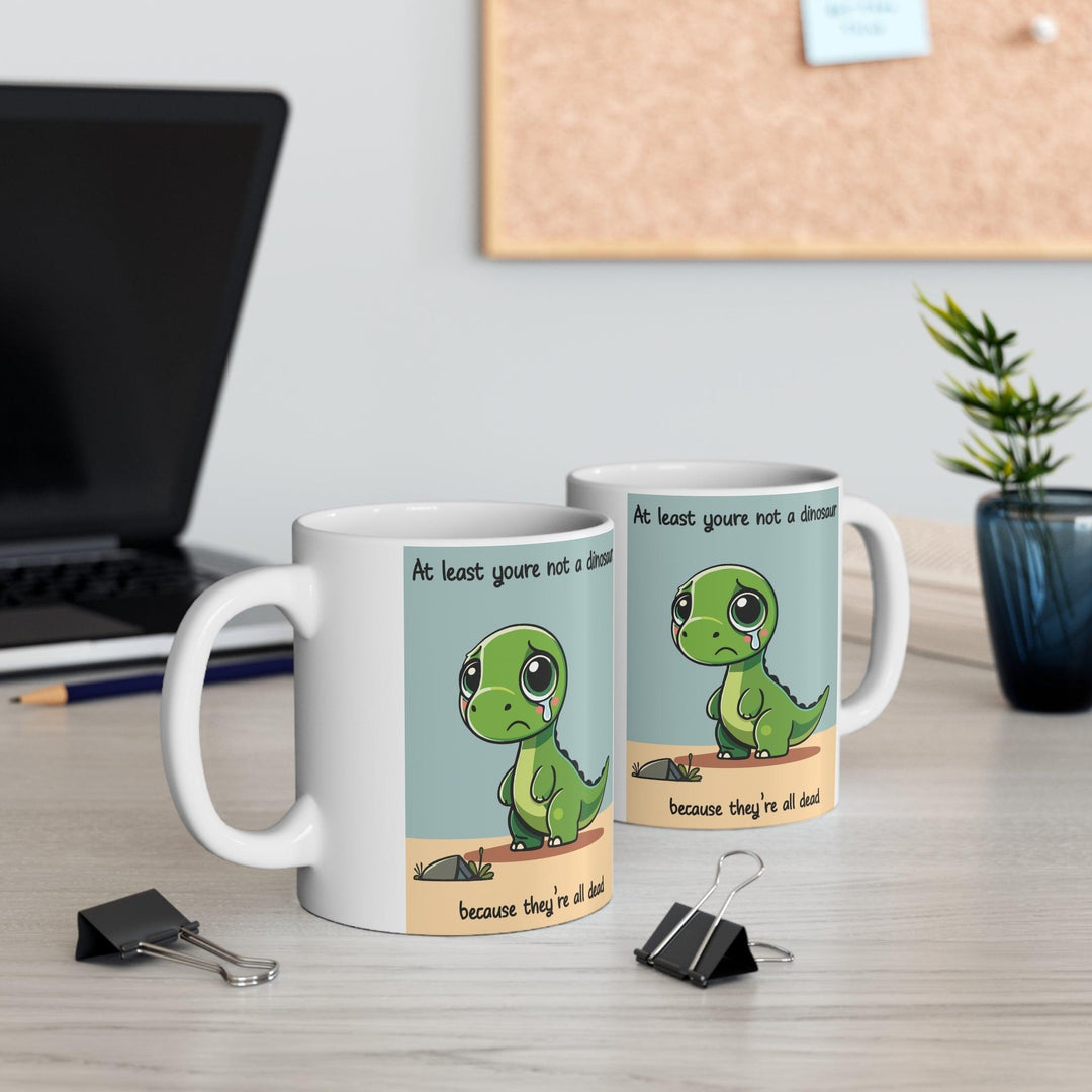 White ceramic mug with a cute dinosaur illustration and the humorous text 'At least you’re not a dinosaur,' adding fun to any morning routine.