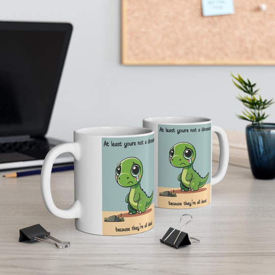White ceramic mug with a cute dinosaur illustration and the humorous text 'At least you’re not a dinosaur,' adding fun to any morning routine.