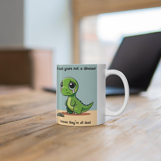 White ceramic mug with a cute dinosaur illustration and the humorous text 'At least you’re not a dinosaur,' adding fun to any morning routine.
