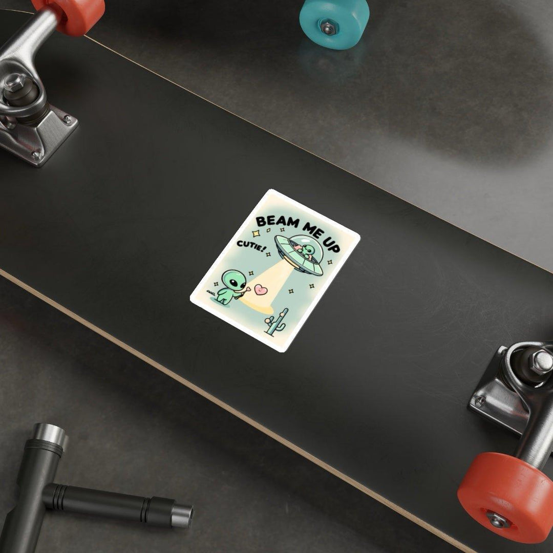A close-up view of a skateboard deck showcases a "Beam Me Up, Cutie" sticker from  KEOLIX , featuring an adorable alien in a UFO lifting a cactus into space. The charming sticker displays the words "BEAM ME UP" and "CUTE!" encircled by hearts. Set on a gray floor with tools around, this scene invites you on an interstellar journey.