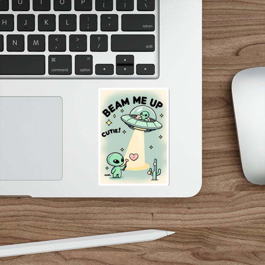 A sticker from  KEOLIX "Beam Me Up, Cutie" collection playfully adorns a laptop beside a mouse, featuring a cartoon alien being whisked away by a UFO. The text proudly declares "BEAM ME UP CUTIE!" amidst twinkling hearts and stars, with a charming cactus adding to this whimsical cosmic scene.