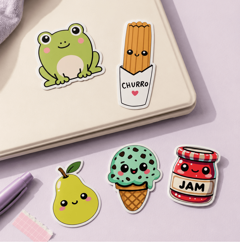 Four cute cartoon stickers arranged on a pale lavender surface next to a cream notebook. The stickers depict a green frog, a churro in a paper bag labeled 'CHURRO', a green ice cream scoop with black polka dots in a waffle cone labeled 'ICE', and a red jam jar labeled 'JAM'.