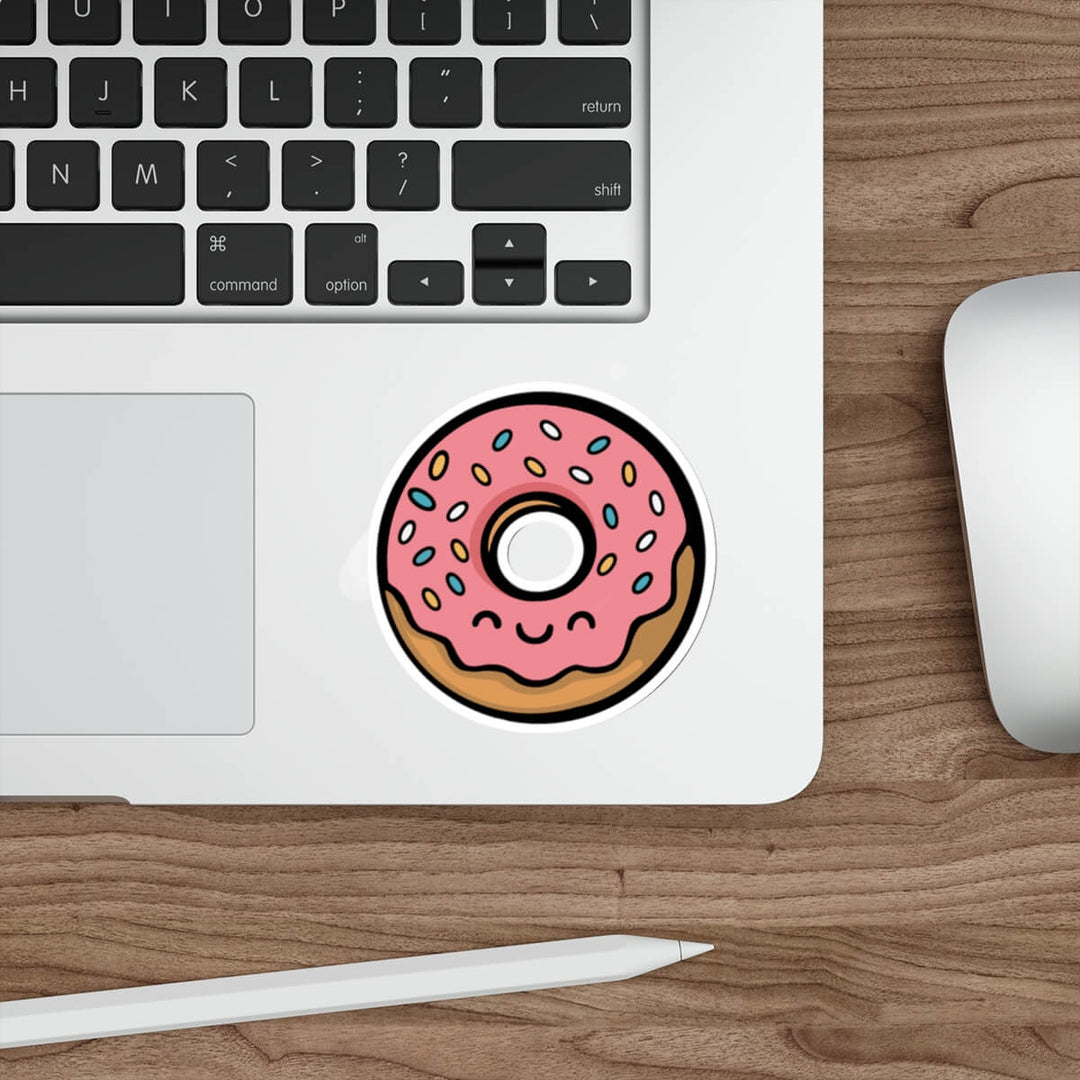 Cute pink frosted donut sticker with rainbow sprinkles and a kawaii smiling face, applied to a silver laptop near a keyboard
