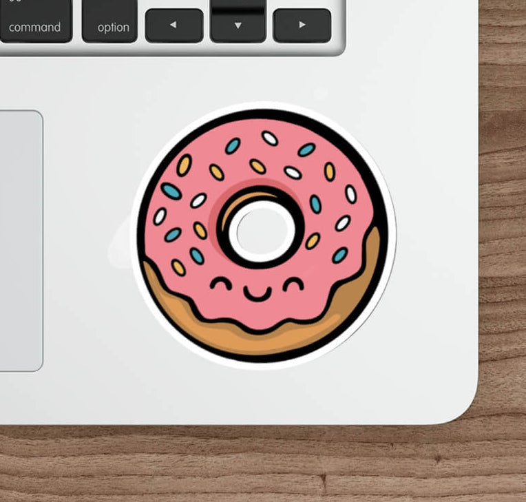 Cute pink frosted donut sticker with rainbow sprinkles and a kawaii smiling face, applied to a silver laptop near a keyboard
