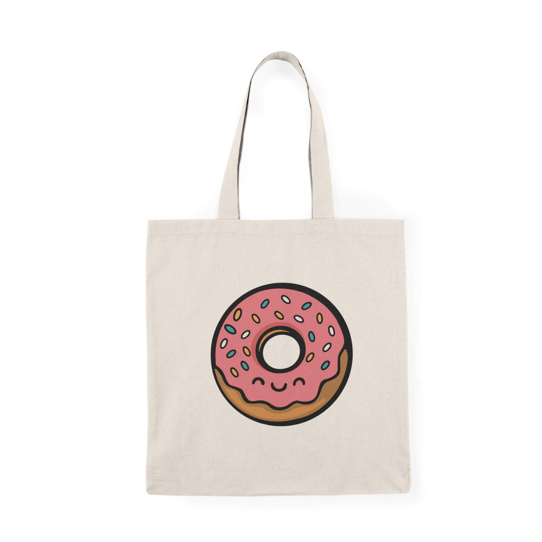 Canvas tote bag with a cute pink frosted donut illustration, complete with sprinkles and a smiling kawaii face, printed on a neutral beige background.
