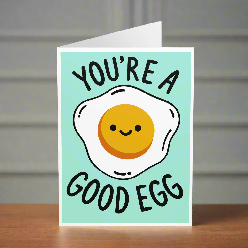 A cute greeting card featuring a smiling fried egg on a pastel teal background with the text "You're a Good Egg" in playful black font.