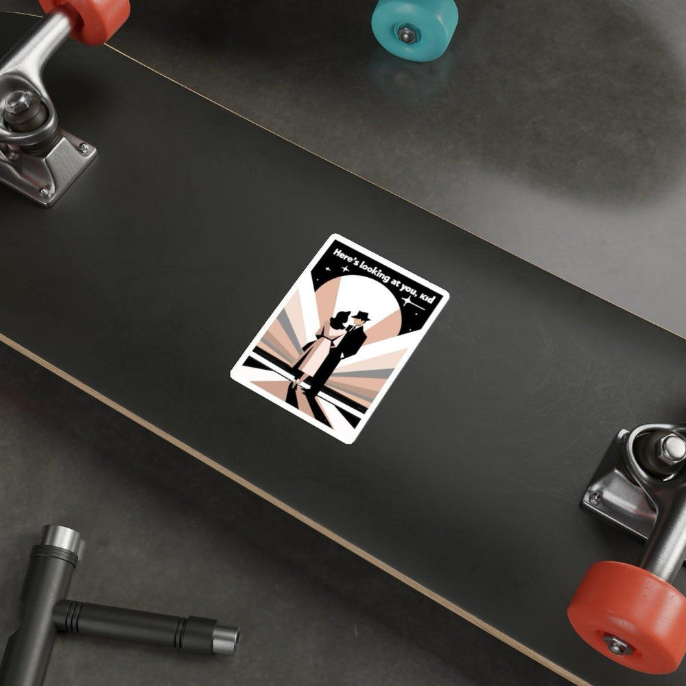 A skateboard rests on the floor, adorned with a Baby Keo's "Here's Looking at You, Kid" waterproof sticker on its deck. The sticker showcases an illustration of two people in an embrace against a stylized sunburst background, with the phrase "Here's looking at you, kid" above them. Various skateboard tools scattered nearby add to the scene's vintage glamour.