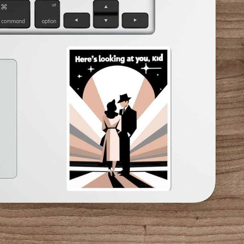 A sticker with an illustration of a romantic scene, placed on a laptop next to a keyboard and a stylus, on a wooden desk.