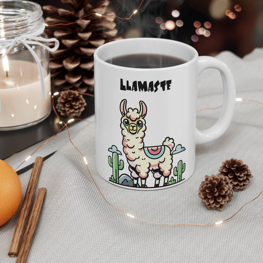 White ceramic mug featuring a charming llama in a yoga pose with the playful text 'Llamaste,' set against a neutral backdrop with cozy pinecones.