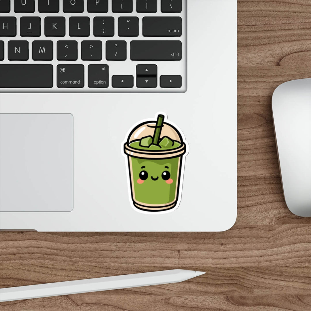 Glossy vinyl sticker of a smiling matcha drink in a dome-lid cup with blush cheeks and a straw, styled on a laptop next to a keyboard and pen.

