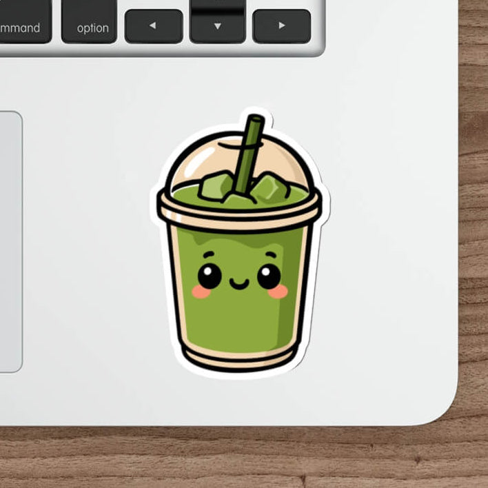 Glossy vinyl sticker of a smiling matcha drink in a dome-lid cup with blush cheeks and a straw, styled on a laptop next to a keyboard and pen.

