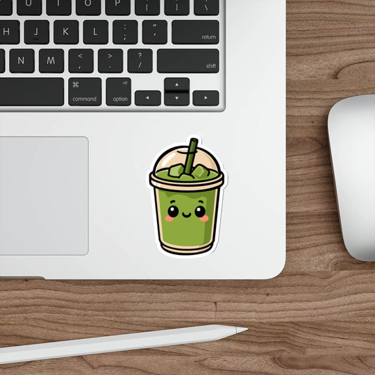 Glossy vinyl sticker of a smiling matcha drink in a dome-lid cup with blush cheeks and a straw, styled on a laptop next to a keyboard and pen.

