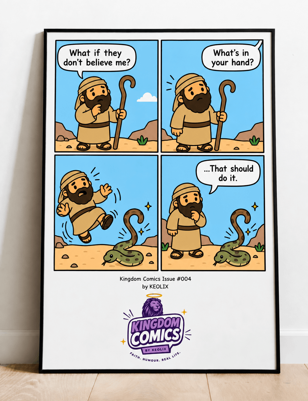 A framed four-panel comic print leaning against a white wall on a light wood floor. The comic shows a cartoon bearded figure in a desert with a staff, speech bubbles, and a green snake, with the text &