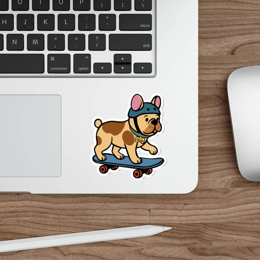 Vinyl sticker of a cartoon French bulldog wearing a helmet and riding a skateboard, placed on a silver laptop beside a white pen and keyboard.