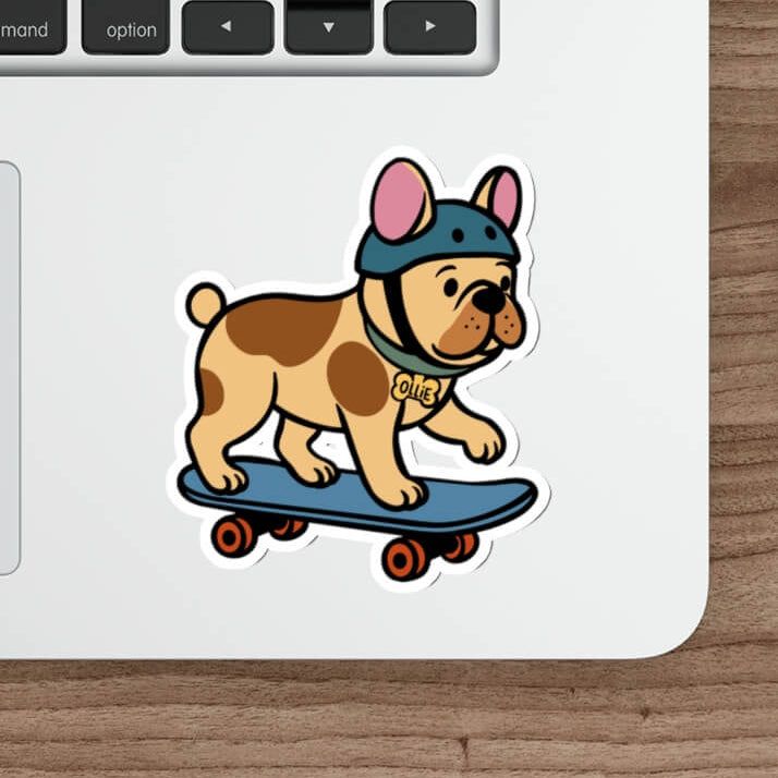 Vinyl sticker of a cartoon French bulldog wearing a helmet and riding a skateboard, placed on a silver laptop beside a white pen and keyboard.