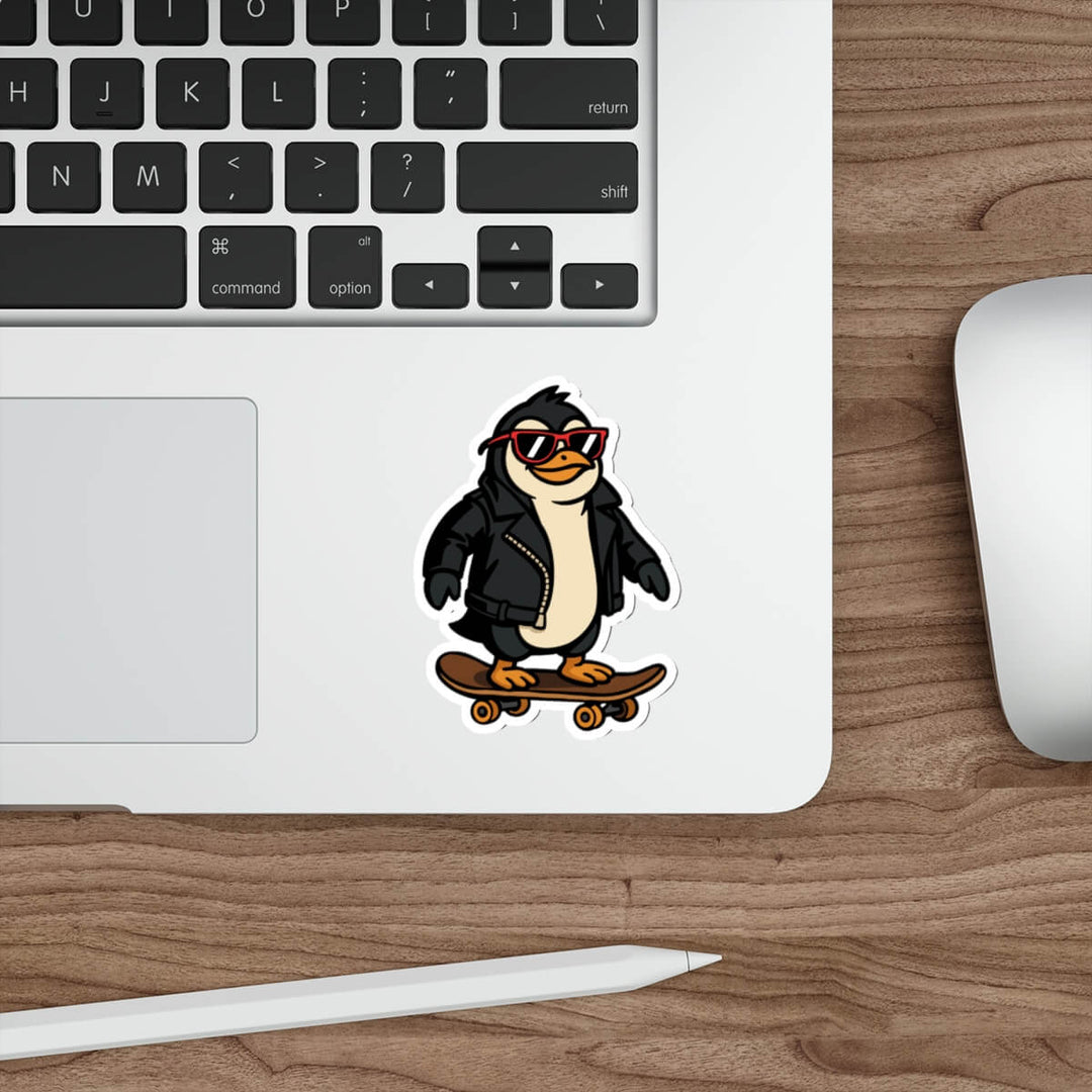 Sticker of a cartoon penguin in a black leather jacket and red sunglasses riding a skateboard, placed on a laptop next to a pencil.