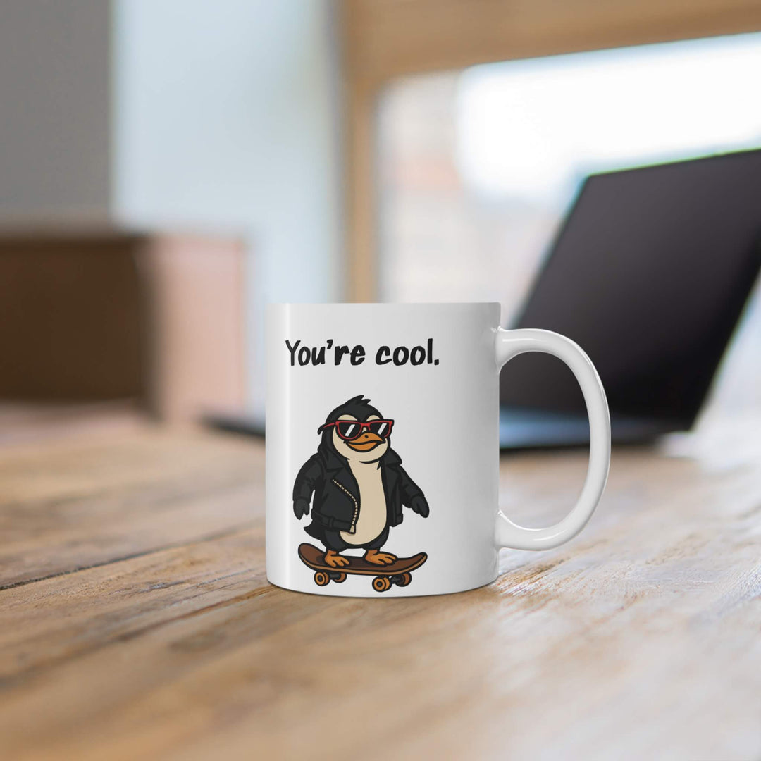 White ceramic mug with the phrase “You’re cool.” above a cartoon penguin wearing sunglasses and a leather jacket, riding a skateboard.

