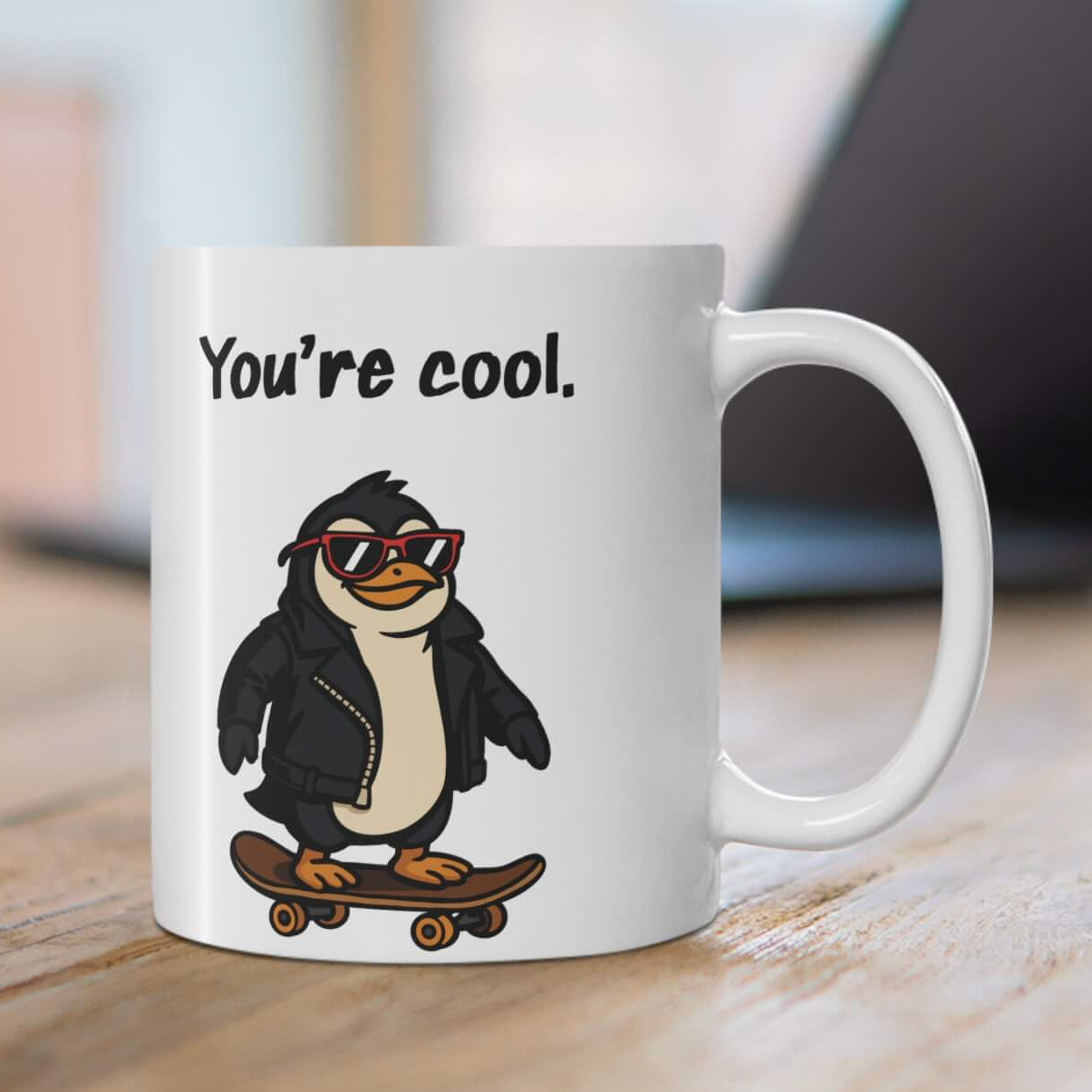 White ceramic mug with the phrase “You’re cool.” above a cartoon penguin wearing sunglasses and a leather jacket, riding a skateboard.

