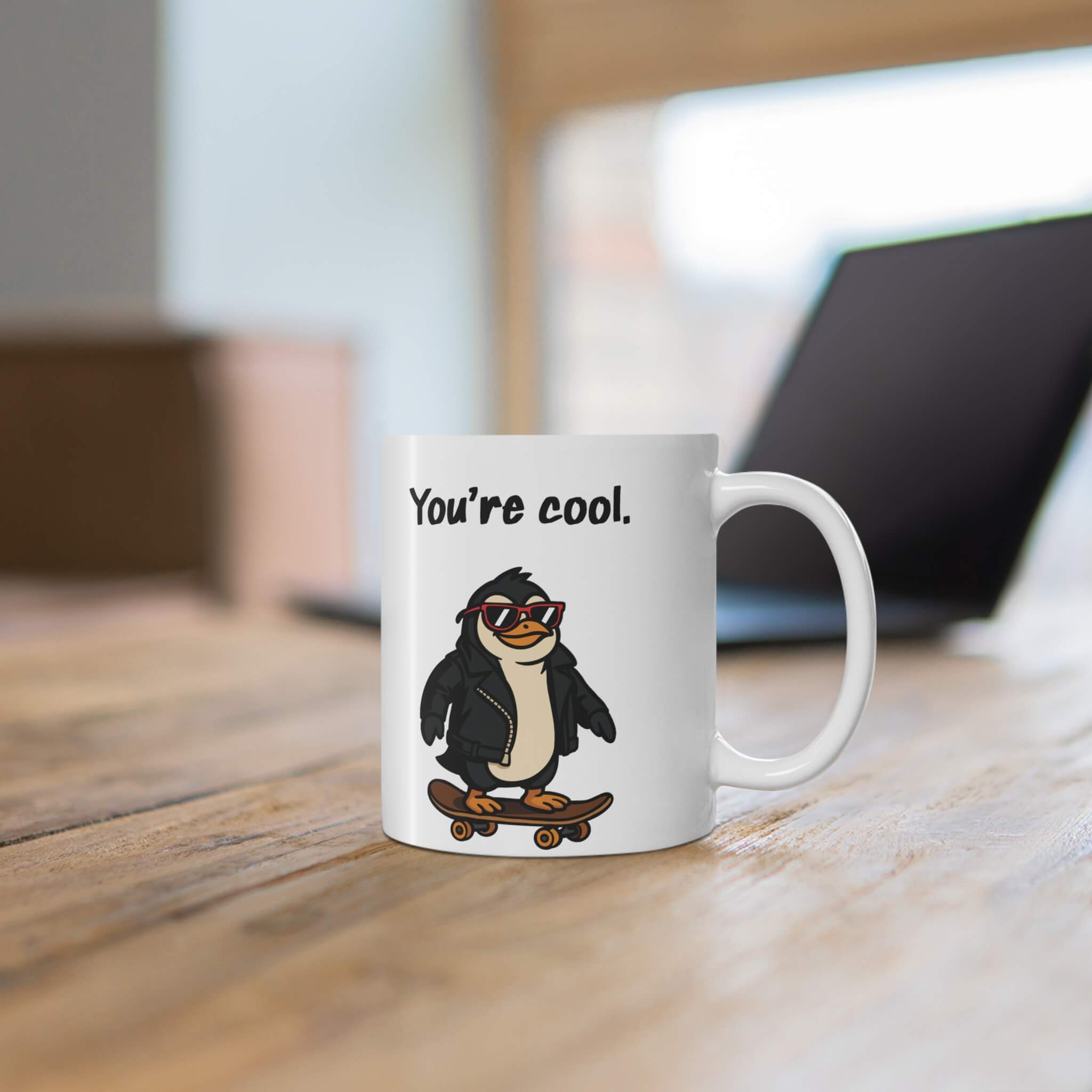 White ceramic mug with the phrase “You’re cool.” above a cartoon penguin wearing sunglasses and a leather jacket, riding a skateboard.

